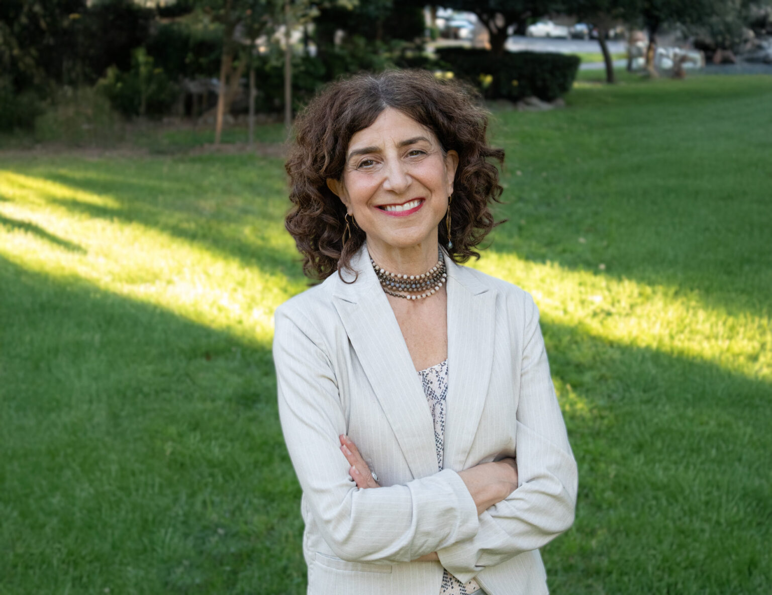 portrait of lori varlotta, standing outside in a white blazer on a green background of grass featuring dappled lighting.