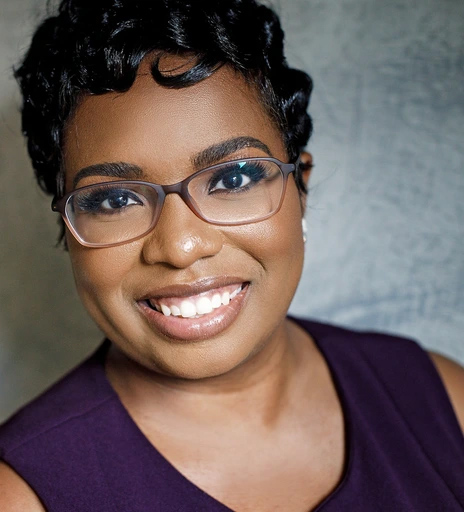Headshot of a smiling person with short dark hair wearing glasses and a sleeveless dark purple top, looking directly at the camera against a softly blurred gray background.