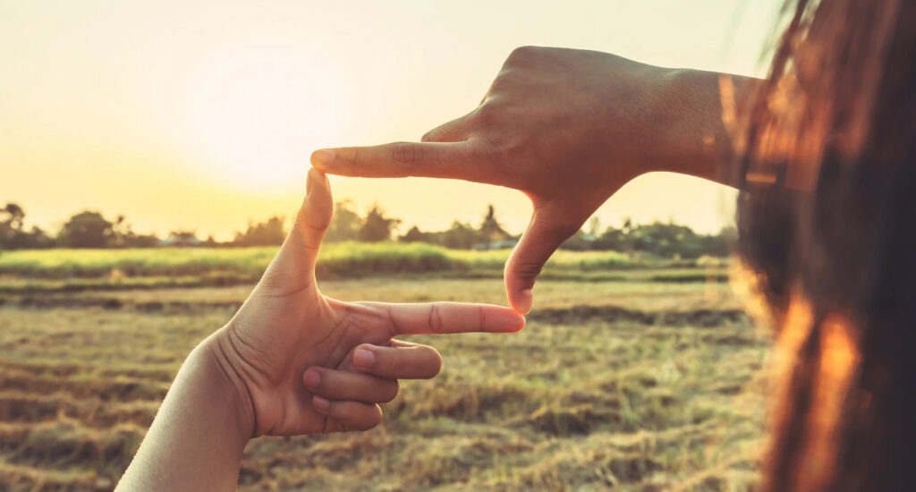 Close-up of a person using both hands to create a rectangular frame while looking across an open field at sunset, symbolizing perspective, vision, or seeing possibilities.