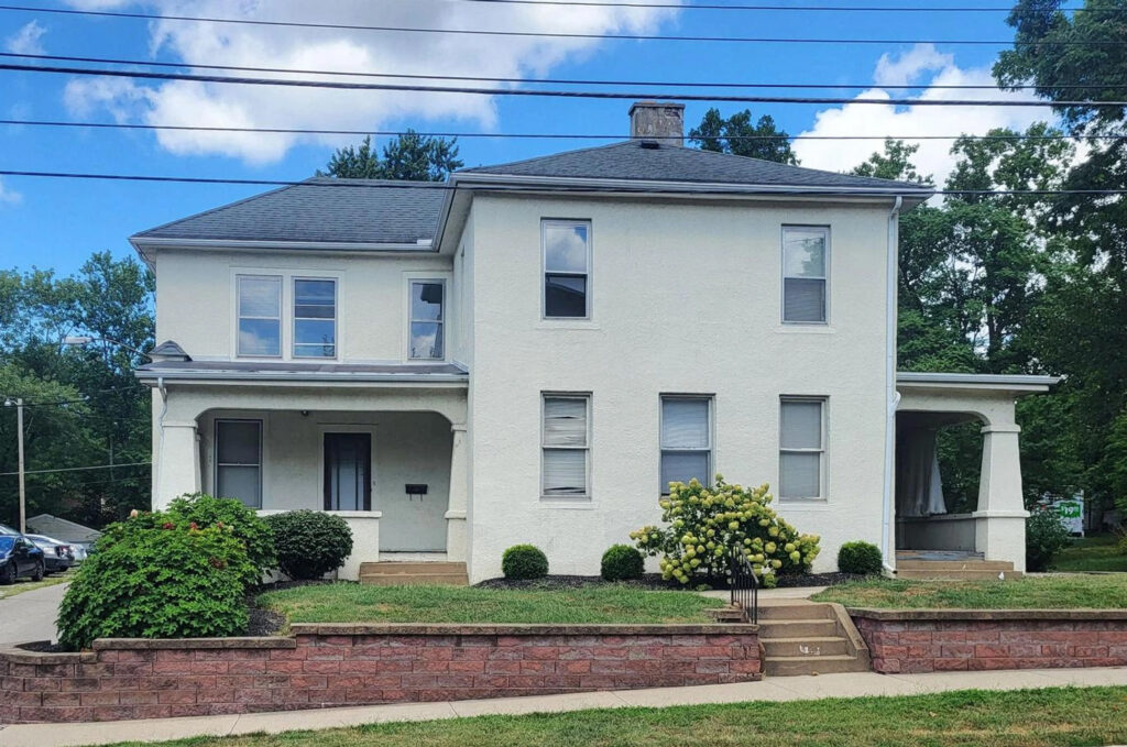 Front view of a large two-story white stucco house with a dark gray roof, two covered front porches, and multiple rectangular windows. A low red brick retaining wall borders the front lawn, with neatly trimmed shrubs and a blooming hydrangea along the foundation. Concrete steps lead from the sidewalk to the entrance under a bright blue sky with scattered clouds.