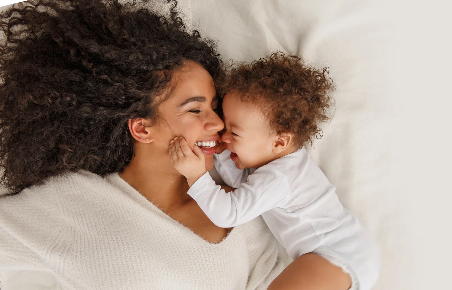 A smiling parent and young child lie face-to-face on a bed, laughing as the child gently cups the parent's cheeks. Both wear white clothing, creating a warm, affectionate moment against soft white bedding.