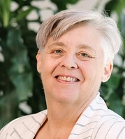 Sue Constable-Headshot of a smiling person with short gray hair wearing a white pinstriped blazer over a light top, facing the camera with leafy greenery softly blurred in the background.