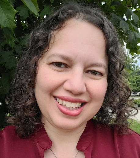 Syntia Santos Diez, PhD, NCC- Smiling person with shoulder-length dark curly hair wearing a burgundy blouse, photographed outdoors beneath leafy green trees. The person is looking directly at the camera with a warm expression, and the background is softly blurred with greenery and a sidewalk.
