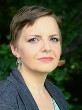 Erin Berzins, Ph.D., LMHC - Director of Clinical Training, CMHC Low-Residency Program; Clinical Mental Health Counseling; Graduate School of Counseling, Psychology & Therapy, Headshot of a person with short brown hair wearing a dark gray blazer and light-colored top, looking toward the camera with a slight smile against a softly blurred green outdoor background.